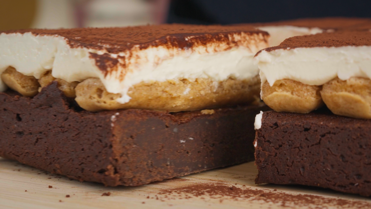 Close-up of layered chocolate brownie dessert with thick white cream and biscotti pieces, dusted with cocoa powder, on a wooden surface.